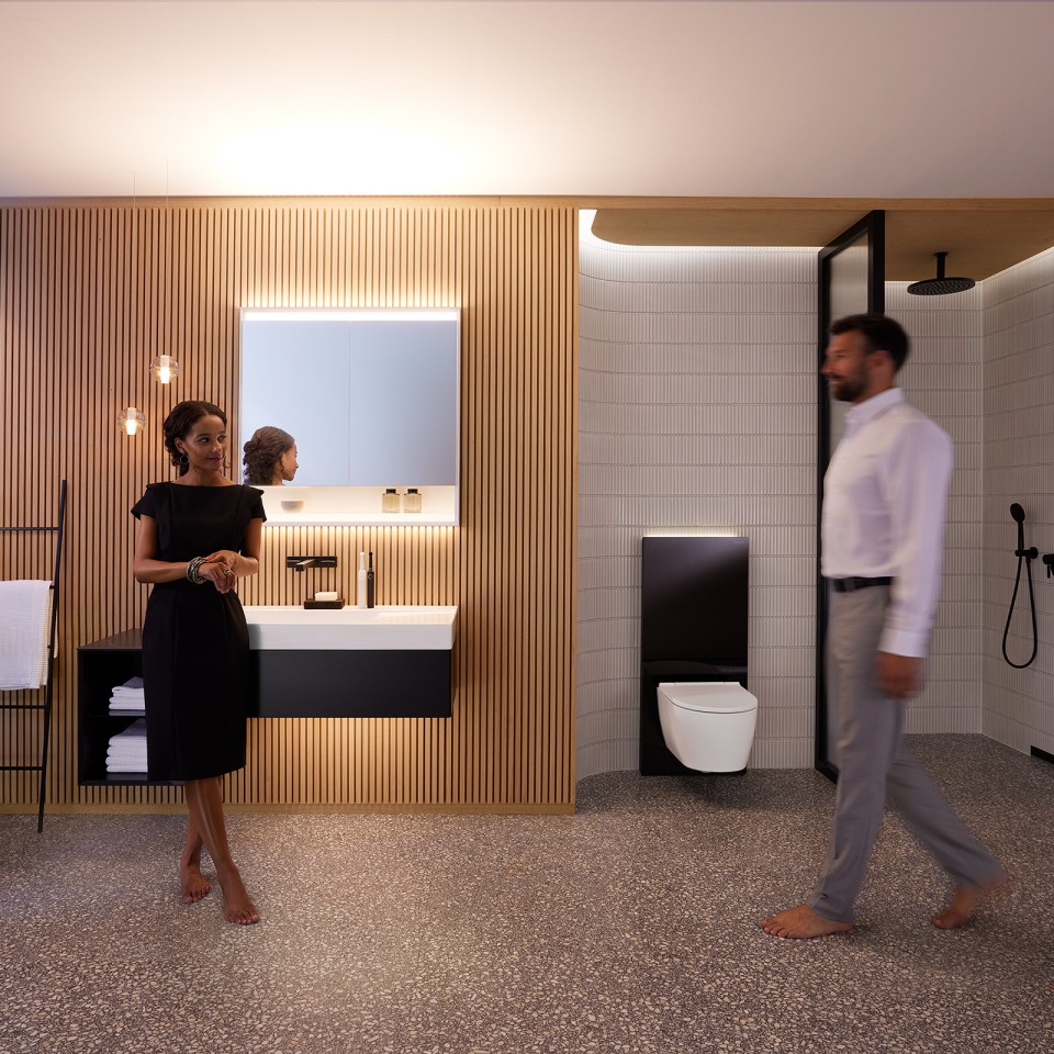 Homme et femme dans une salle de bains parfaitement éclairée avec les meubles de salle de bains et les céramiques sanitaires Geberit ONE (© Geberit)